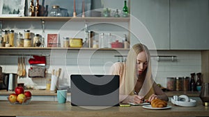 Girl student studying online sitting at kitchen with laptop. Woman making notes.