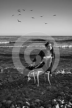 Girl in stylish formal suit, shirt and hat sitting on white chair in water of ocean.