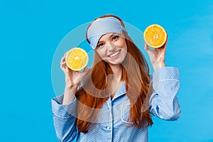 Girl suggesting fruit for breakfast. Cute and tender redhead woman in sleep mask, pyjama, tilt head and holding two
