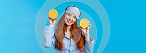 Girl suggesting fruit for breakfast. Cute and tender redhead woman in sleep mask, pyjama, tilt head and holding two