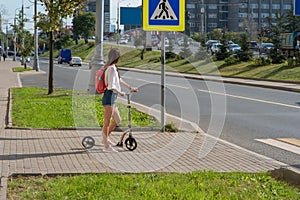 A girl in the summer in the city crosses the road with a scooter, casual wear denim shorts, a white shirt and a pink