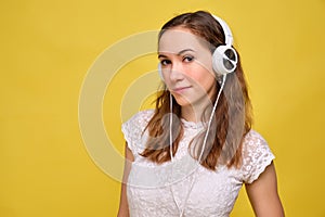 A girl in summer clothes stands on a yellow background, listens to music in white headphones and looks at the camera. Portrait