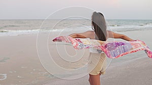 A girl in a summer T-shirt and shorts is walking along the beach looking at the horizon and holding a shawl in her hands