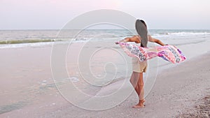 A girl in a summer T-shirt and shorts is walking along the beach looking at the horizon and holding a shawl in her hands