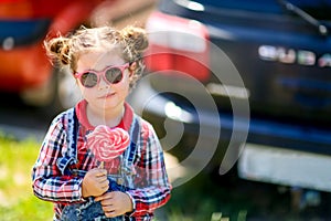 Girl in sunglasses and candy on a stick in her hands on the background of cars