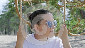 A girl in sunglasses is chilling on a rope swing. Close-up, zoom out