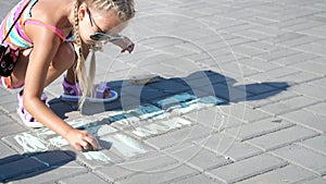 A girl in sunglasses, draws drawings with colored crayons on the asphalt, street tiles. A hot summer day.