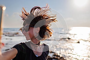 girl in sunglasses with hair flying in the wind, taking a selfie by the sea