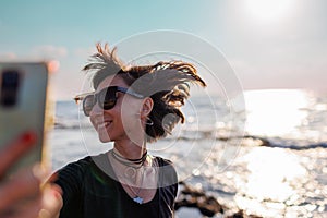 girl in sunglasses with hair flying in the wind, taking a selfie by the sea