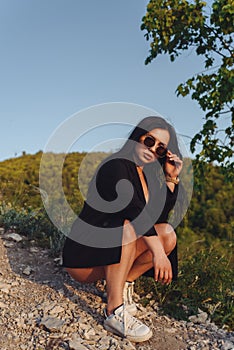 The girl in sunglasses is on the hill. The cool brunette is hunkering. The girl is against the sky and greenery