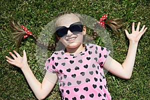 Girl in sunglasses lie on green grass, dressed in pink clothes with hearts, bright sun, summer outdoor, top view