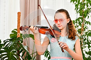 A girl in sunglasses plays the violin at home among green plants