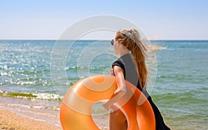 A girl on a sunny day stands on the seashore with an inflatable ring and looks thoughtfully into the distance.