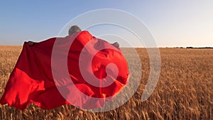 Girl super hero running across field with wheat in red cloak against blue sky.