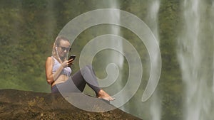 Girl surfs internet sitting on stone by waterfall