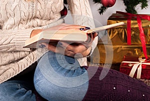 Girl in sweater holding book in hands and Christmas tree and gift box in background