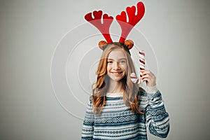 Girl in a sweater with red horns smiles, holding a Christmas lollipop in her hand. She will be happy to decorate the