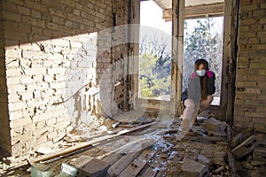 Girl in a sweatshirt and brown boots is sittig in an abandoned building in a blue medical mask and pink gloves