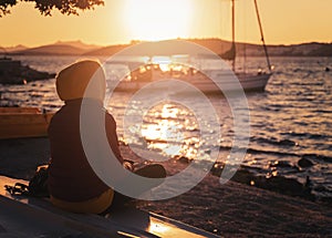 A girl in a sweatshirt looks at the sunset on the sea and at the boats on the shore in the sunshine