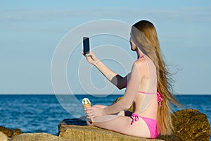 Girl in swimsuit with ice cream in hand on rocky beach makes selfie phone against the sea