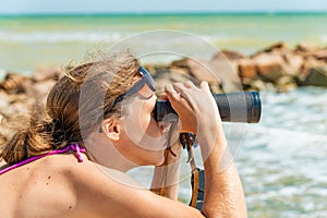 A girl in a swimsuit looks through binoculars at the sea on the beach