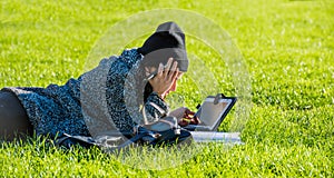 Girl with tablet, lying on green grass and writing on tablet bores from homework