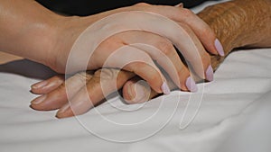 Girl takes and gently touches hand of sick mature woman lying in bed hospital. Granddaughter comforts wrinkled arm of