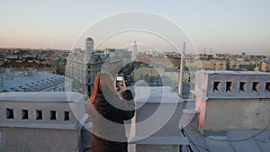 Girl takes photo video on phone stand on terrace with historical buildings view.