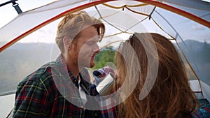 Girl taking care about guy hair in camping tent. Couple drinking tea from mugs