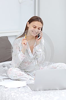 Girl talking on the phone in home. Woman working, learns and using laptop computer in the bedroom. Freelancer. Writing, typing.