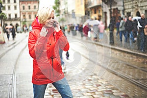 Girl talking on the phone under rain on the street . Portrait of pretty smiling young woman in red bright raincoat