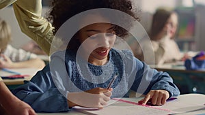 Girl talking with teacher in classroom. Smiling schoolgirl writing in notebook