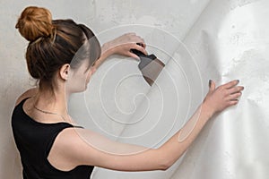 The girl tears off the old wallpaper from the concrete wall and holds a spatula.