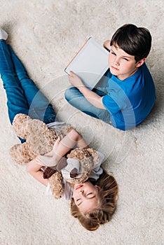 Girl with teddy bear and boy with digital tablet on carpet at home