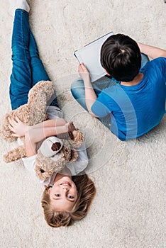 Girl with teddy bear and boy with digital tablet on carpet at home