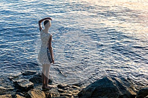 Girl teenager in a dress stands on the seashore, looking into the distance
