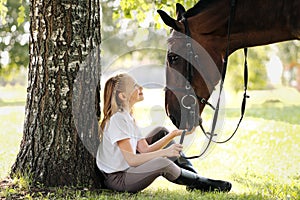 Girl teenager jockey sits in a green clearing under a tree. Feeds a horse an apple and strokes it.