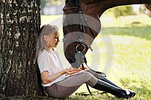 Girl teenager jockey sits in a green clearing under a tree. Feeds a horse an apple and strokes it.