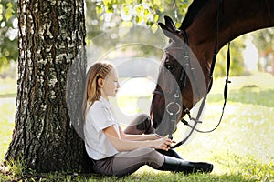 Girl teenager jockey sits in a green clearing under a tree. Feeds a horse an apple and strokes it.