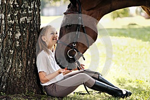 Girl teenager jockey sits in a green clearing under a tree. Feeds a horse an apple and strokes it.