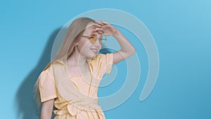 Girl teenager looking far away on blue background in studio. Young girl in yellow glasses and dress peering into