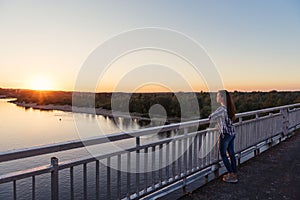 Girl teenager in plaid shirt and jeans stands on a motu on the river with a view of the sunset