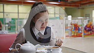 Girl teenager in a school uniform sitting at a table in the school cafeteria eating cakes and drinking tea.