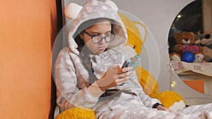 Girl teenager uses a smart phone and eating a tangerine while lying on the bed in warm pajamas.