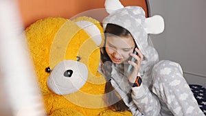 Girl teenager warm pajamas talking on a cell phone with a friend while sitting in bed with a Teddy bear.