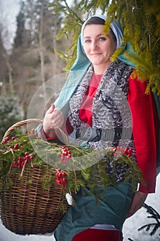 Girl in thick coat and a red sash with basket of fir branches and berries in cold winter day in forest. Medieval peasant