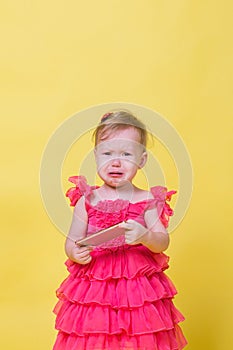 Girl toddler in a pink dress on a yellow background holding a smartphone and crying