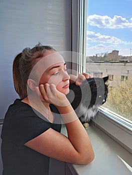 Girl together with cat looking sadly out window staying home during quarantine