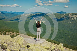 The girl at the top of the mountain raised her hands up. Wide summer mountain view at sunrise and distant mountain range covered.