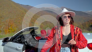 Girl tourist in boho style red jacket dancing near convertible car in mountains.